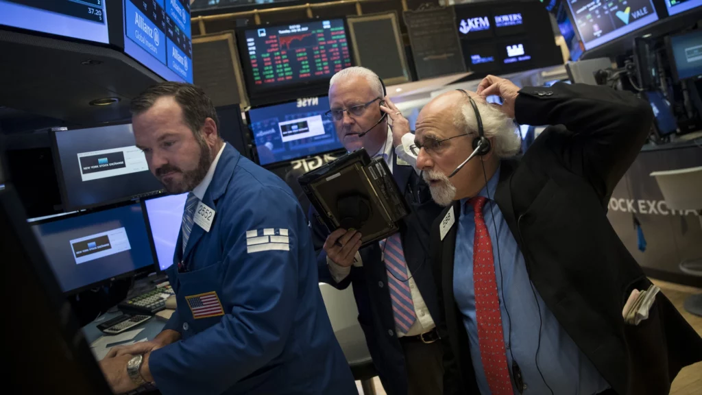 Three New York Stock Exchange traders standing at a trading desk. One trader is scratching his head.